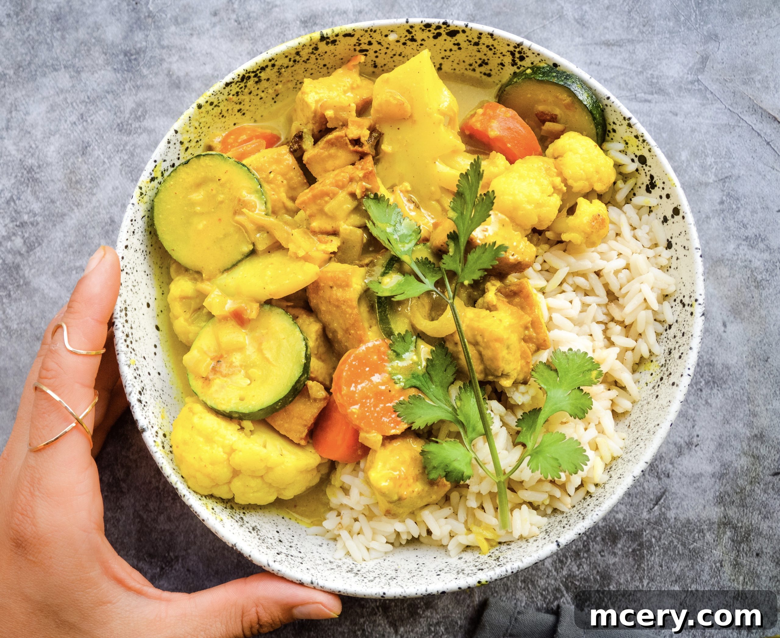 Effortless Tofu and Colorful Vegetable Curry 2 A hearty bowl of Easy Tofu and Vegetable Curry with crispy tofu, zucchini, carrots, and cauliflower, garnished with fresh herbs, served on a gray textured background.
