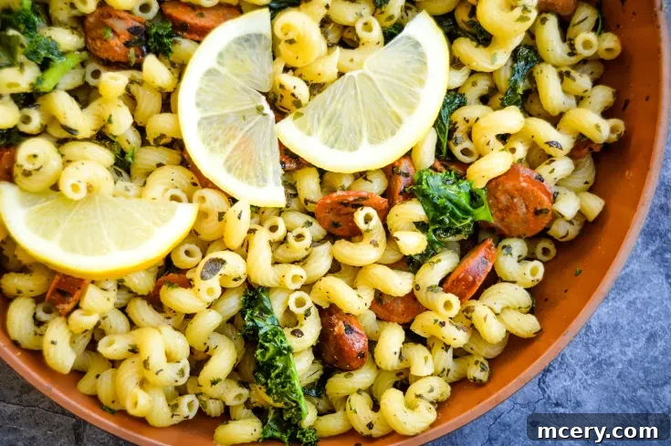 A bowl of Lemon Sausage & Kale Pasta spirals mixed with sautéed kale and sliced sausage, garnished with three lemon wedges on top, placed on a textured, gray surface.