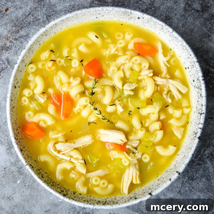 A bowl of chicken noodle soup with macaroni, shredded chicken, carrot slices, and celery in a light yellow broth. The dish is garnished with a sprig of fresh thyme, placed on a textured gray surface.