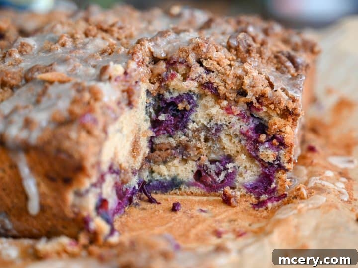 Moist Bakery Style Blueberry Burst Coffee Cake 2 A beautifully baked blueberry coffee cake with a thick streusel topping and a drizzle of glaze, presented on a white plate with a fork.