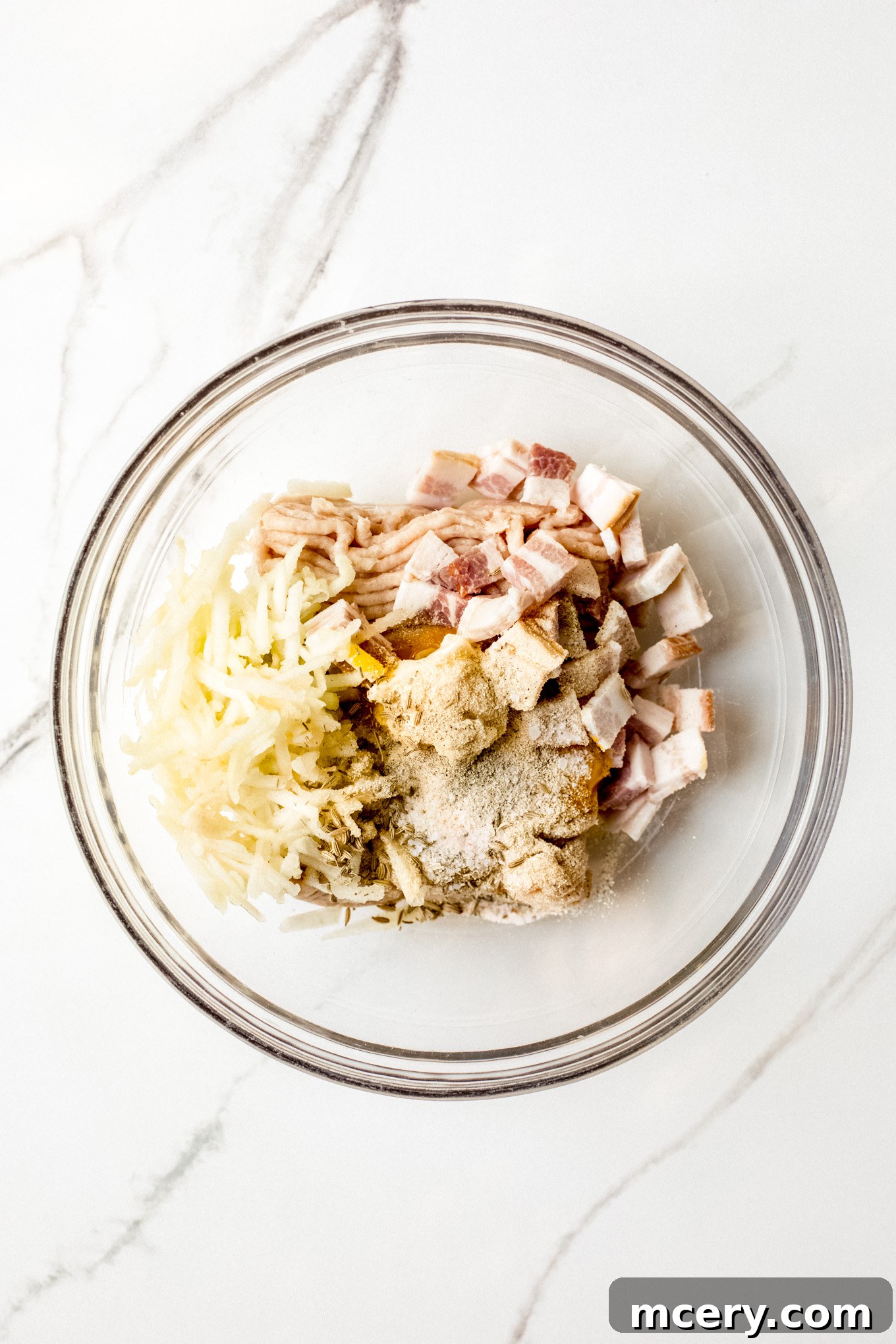 Chicken Apple Sausage Delight 4 A large glass bowl containing ground chicken, finely chopped bacon, shredded apples, and various spices, illustrating the first step of combining ingredients for homemade sausage.