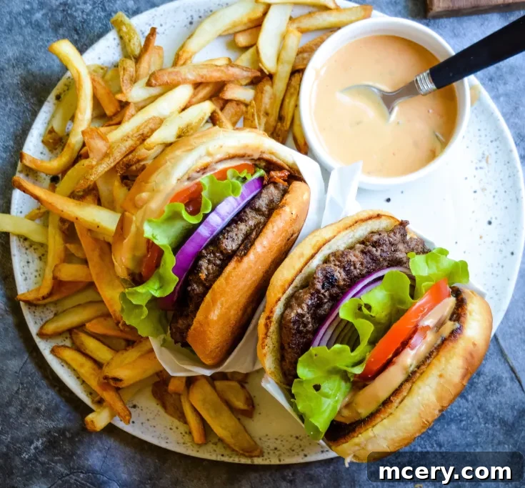 A plate with two burgers filled with lettuce, tomato, pickles, and red onion, surrounded by a generous serving of golden fries. A small bowl of dipping sauce with a spoon is placed on the side.