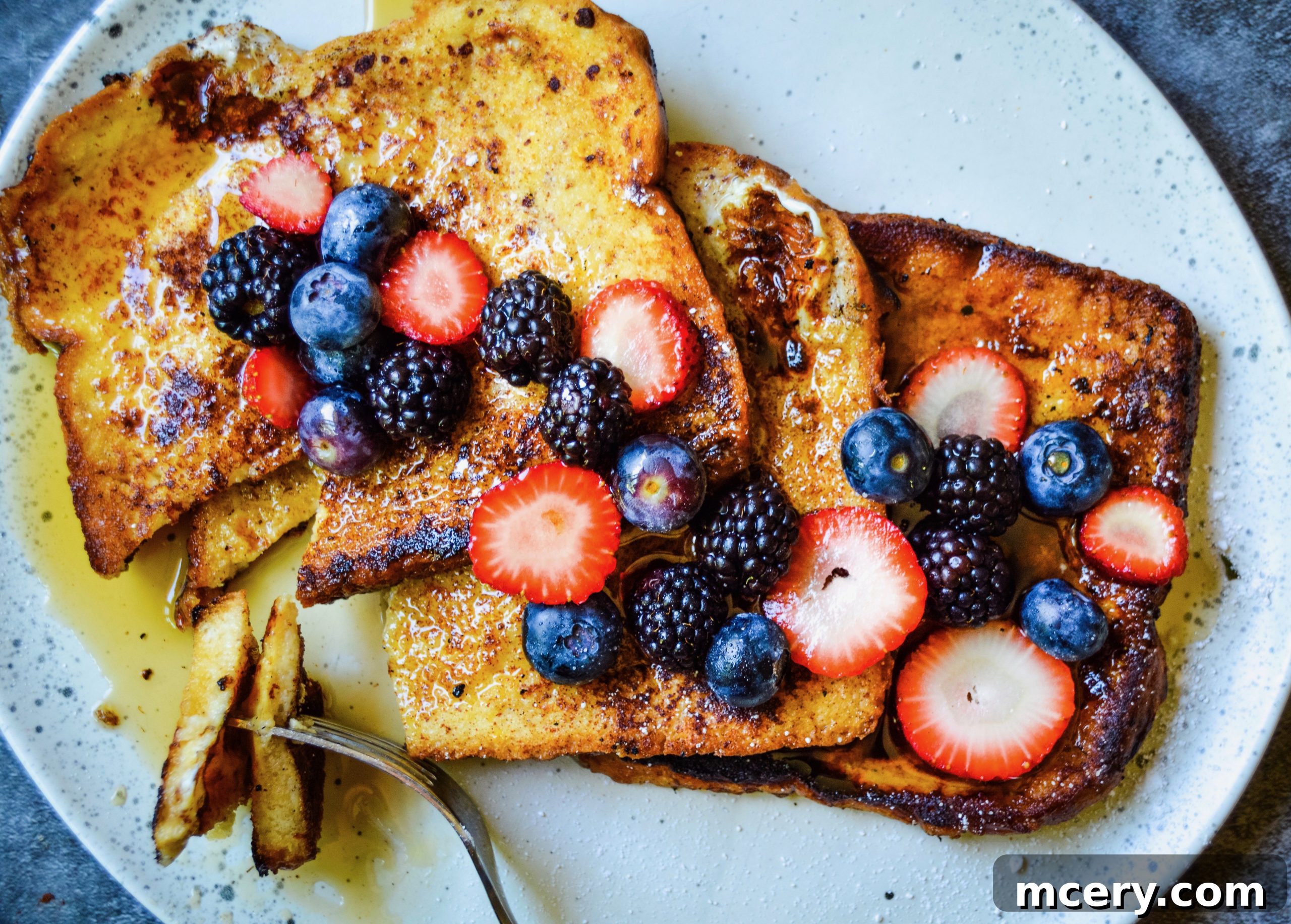 A stack of French Toast slices on a white plate, generously topped with fresh berries and drizzled with maple syrup.