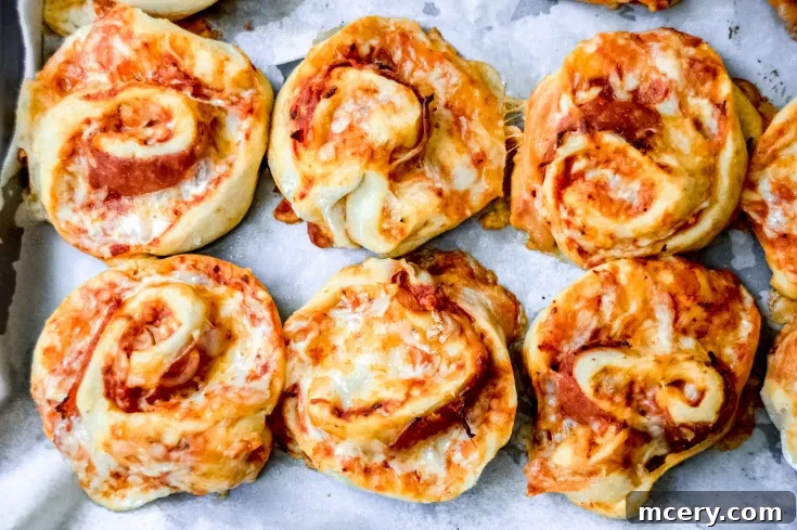 A tray of freshly baked Pizza Pinwheels with Homemade Dough arranged in rows. Each roll has a golden-brown crust with visible swirls of tomato sauce, melted cheese, and hints of pepperoni. They are placed on parchment paper.