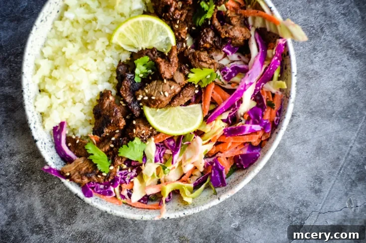 A colorful Asian Steak Bites & Coleslaw Bowl featuring white rice, marinated beef slices, vibrant coleslaw with red cabbage and carrots, garnished with cilantro and lime wedges. The dish is presented in a speckled ceramic bowl.