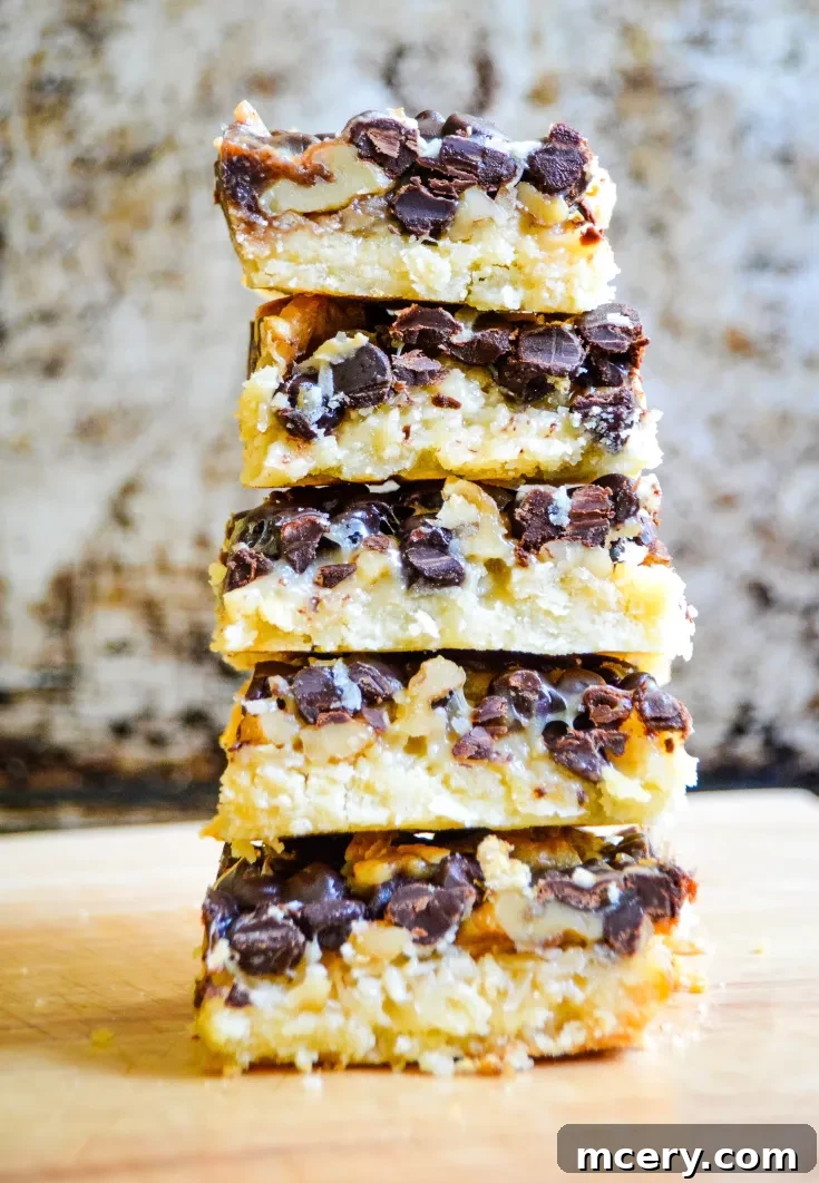 A vertical stack of Magic Cookie Bars with Condensed Coconut Milk sits on a wooden surface. The bars are layered with chocolate chips, coconut, and a creamy filling, showcasing a textured and rich appearance. The background is a rustic, blurred brown and white canvas.