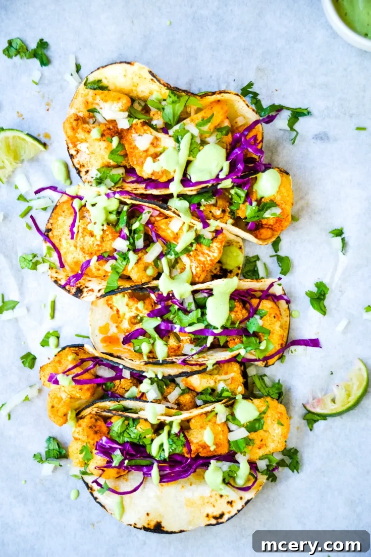 Roasted Cauliflower Tacos filled with crispy cauliflower, purple cabbage, cilantro, and drizzled with green sauce are arranged on parchment paper. Lime wedges are placed nearby for garnish.