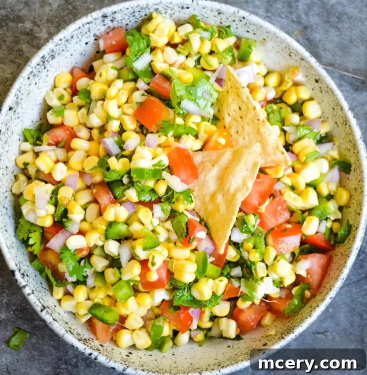 A colorful Fresh Corn Salsa in a speckled bowl features corn, diced tomatoes, chopped onions, jalapeños, cilantro, and a few tortilla chips on top, set against a gray background.