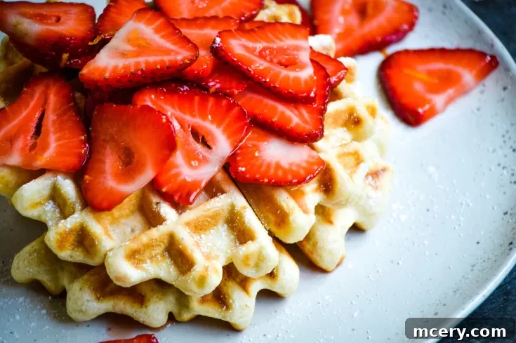 A plate of waffles topped with fresh strawberry slices. The waffles are golden brown and the strawberries are vibrant red, creating a colorful contrast on the white plate. The background is a dark, textured surface.