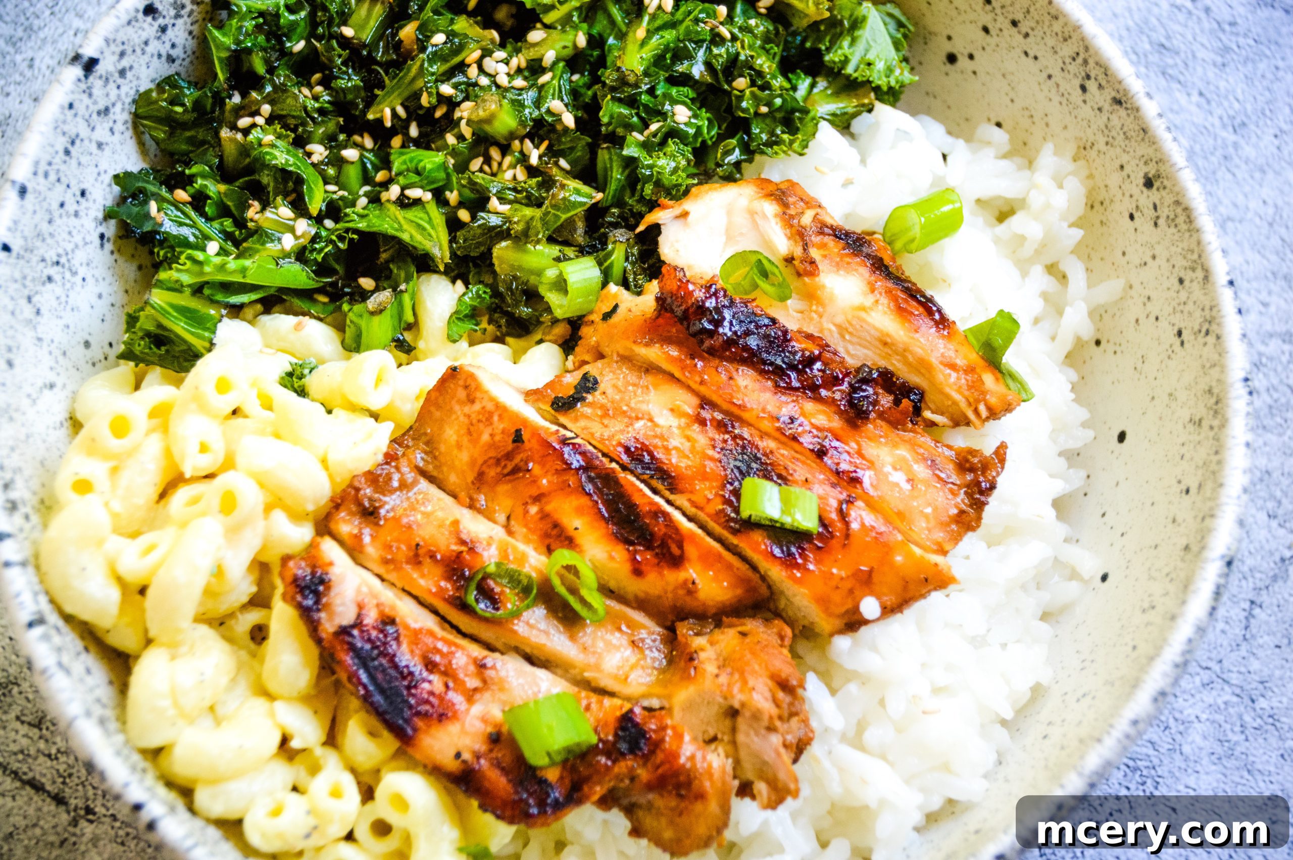 A beautifully presented teriyaki chicken bowl with rice, green onions, and sesame seeds, alongside a side of macaroni salad and steamed vegetables.