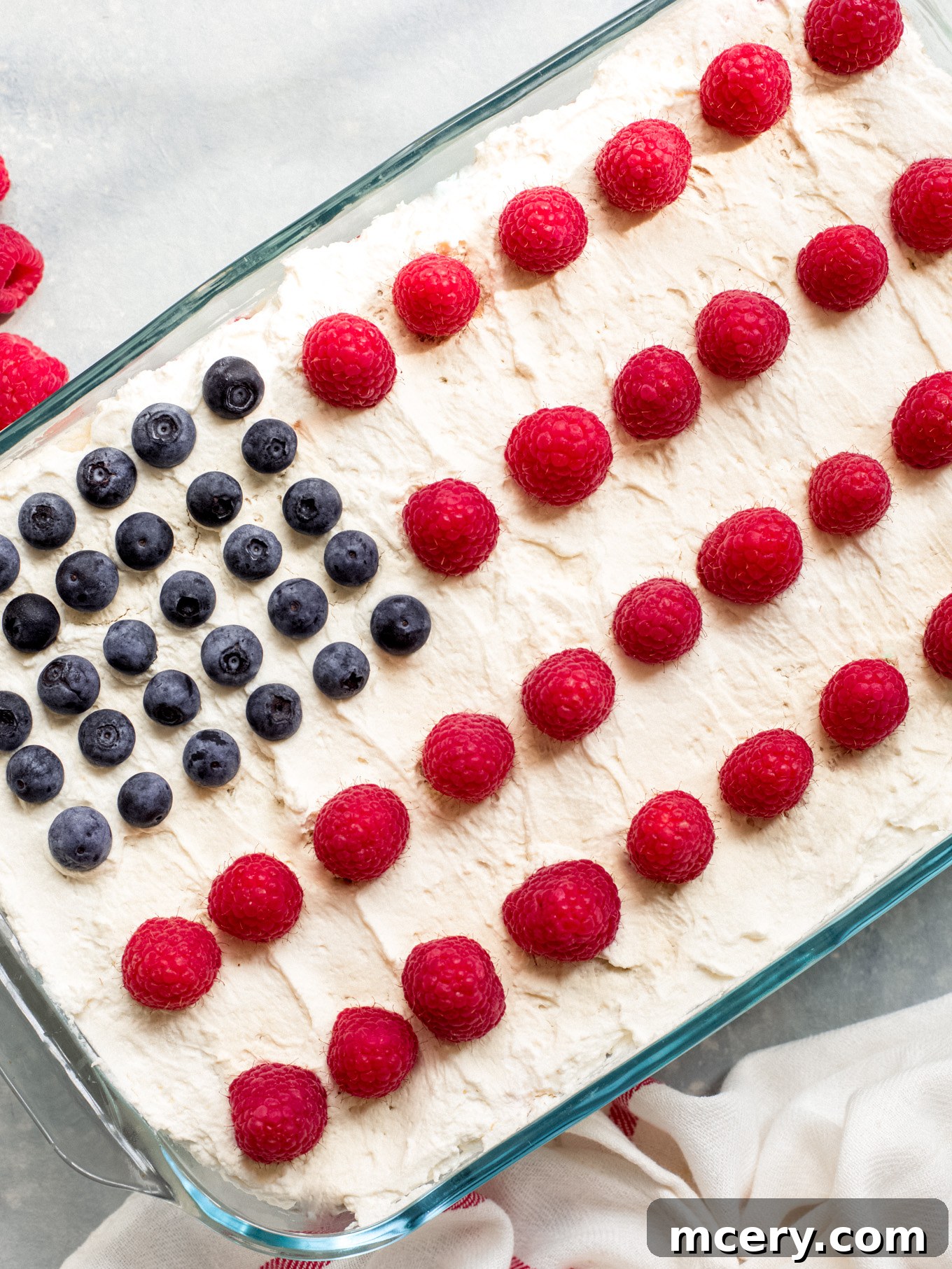 A stunning Icebox Cake in a clear glass dish, beautifully decorated with red strawberries and raspberries for stripes, and blue blueberries for stars, mimicking the American flag. This no-bake summer dessert is perfect for 4th of July celebrations.