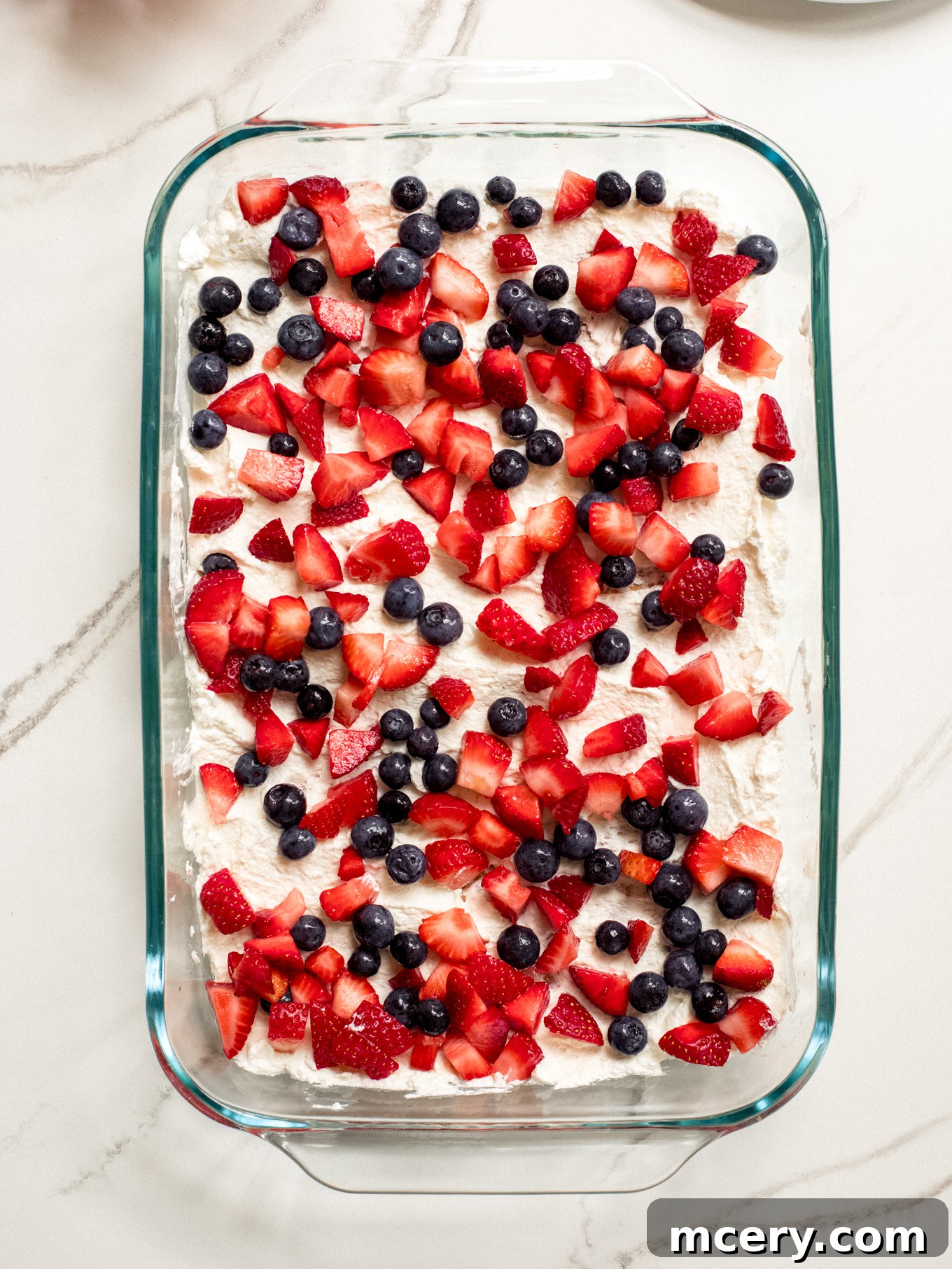 A layer of cream and half of the mixed chopped fruit (strawberries and blueberries) spread over graham crackers in a glass baking dish, forming a colorful layer.