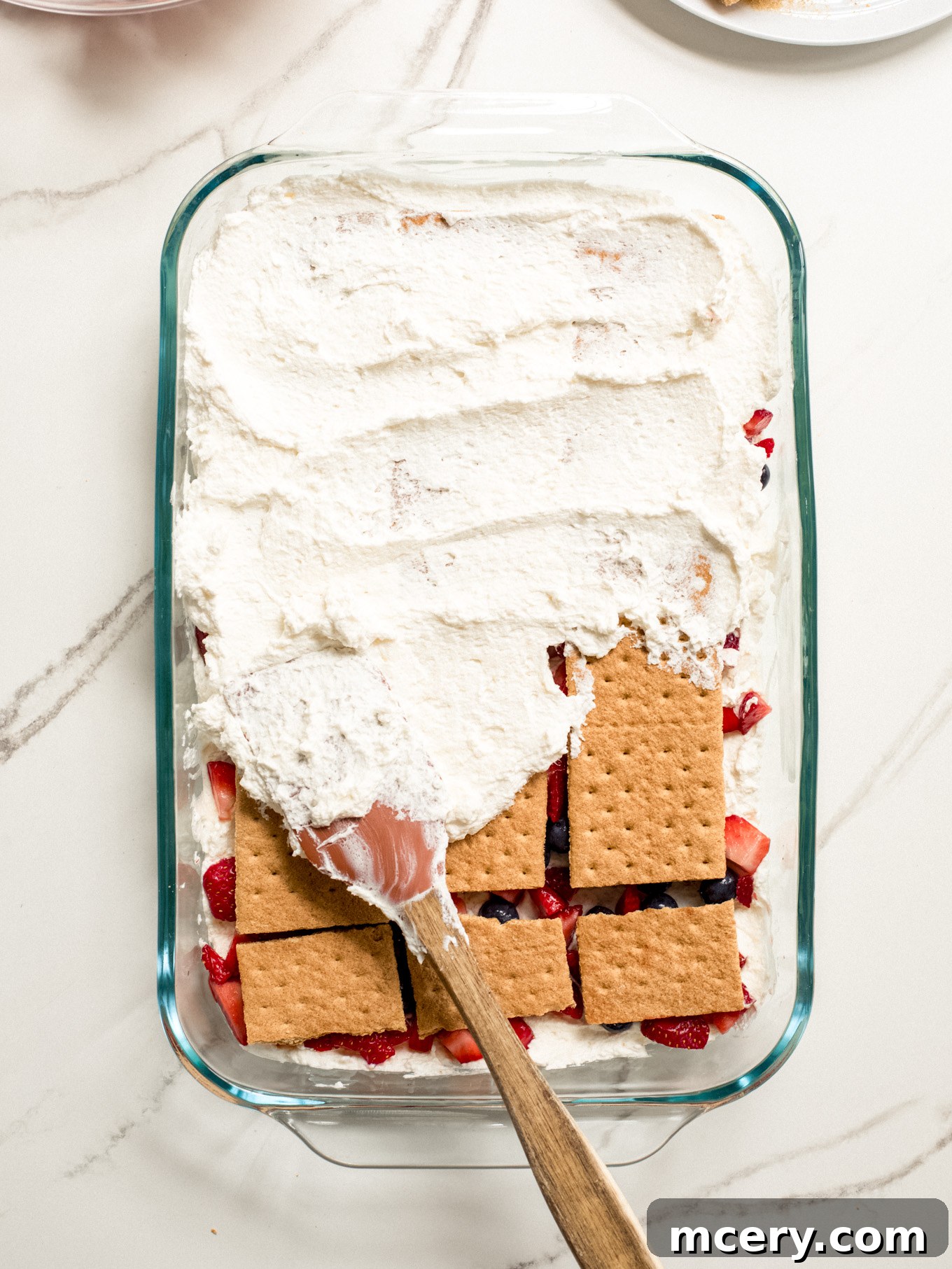 A rubber spatula spreading a generous layer of whipped cream over the graham crackers in the glass baking dish, ensuring even coverage for the next set of layers.