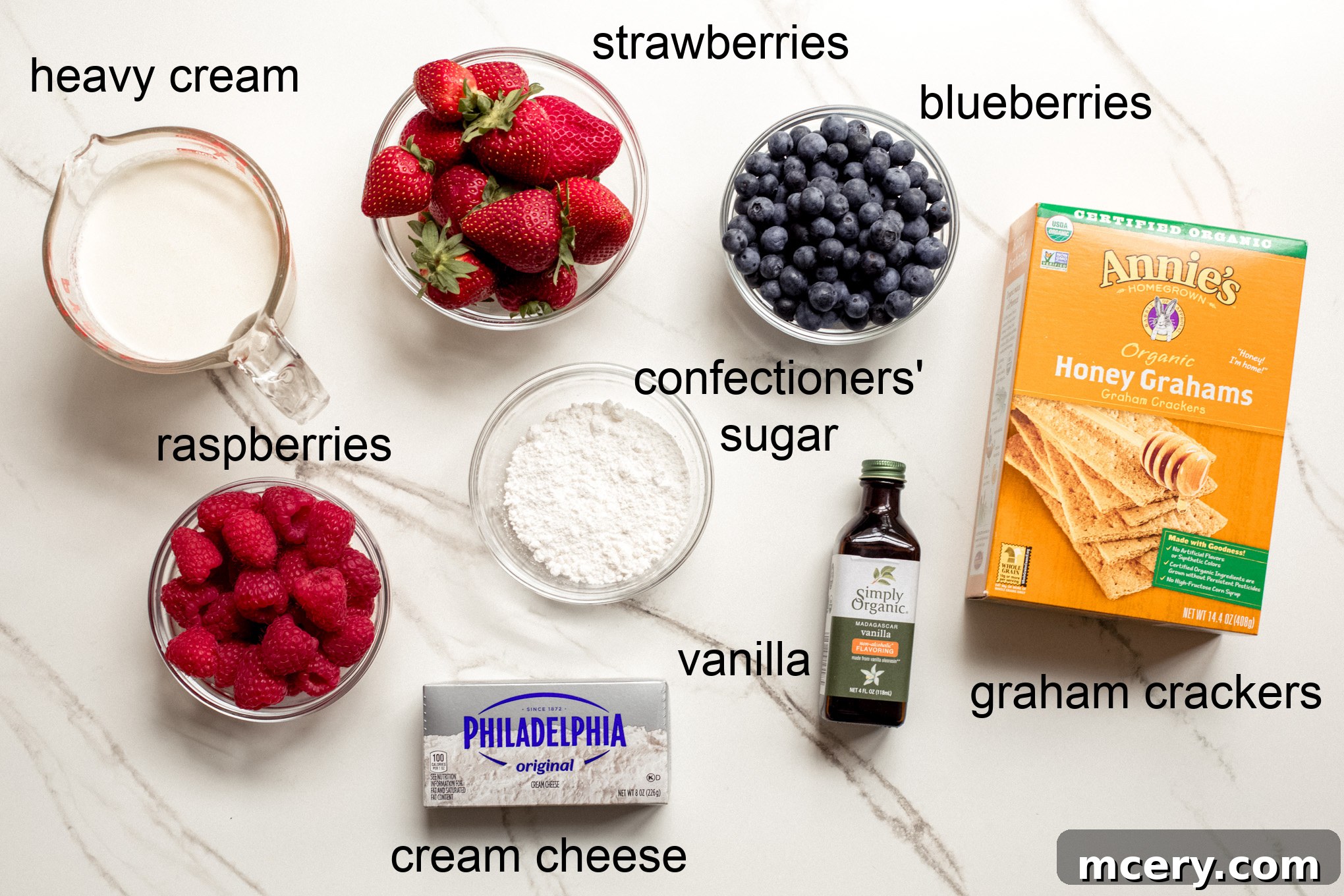 All the fresh ingredients laid out on a white surface for a no-bake icebox cake recipe, including fresh strawberries, blueberries, raspberries, graham crackers, cream cheese, heavy cream, confectioners' sugar, and vanilla extract.