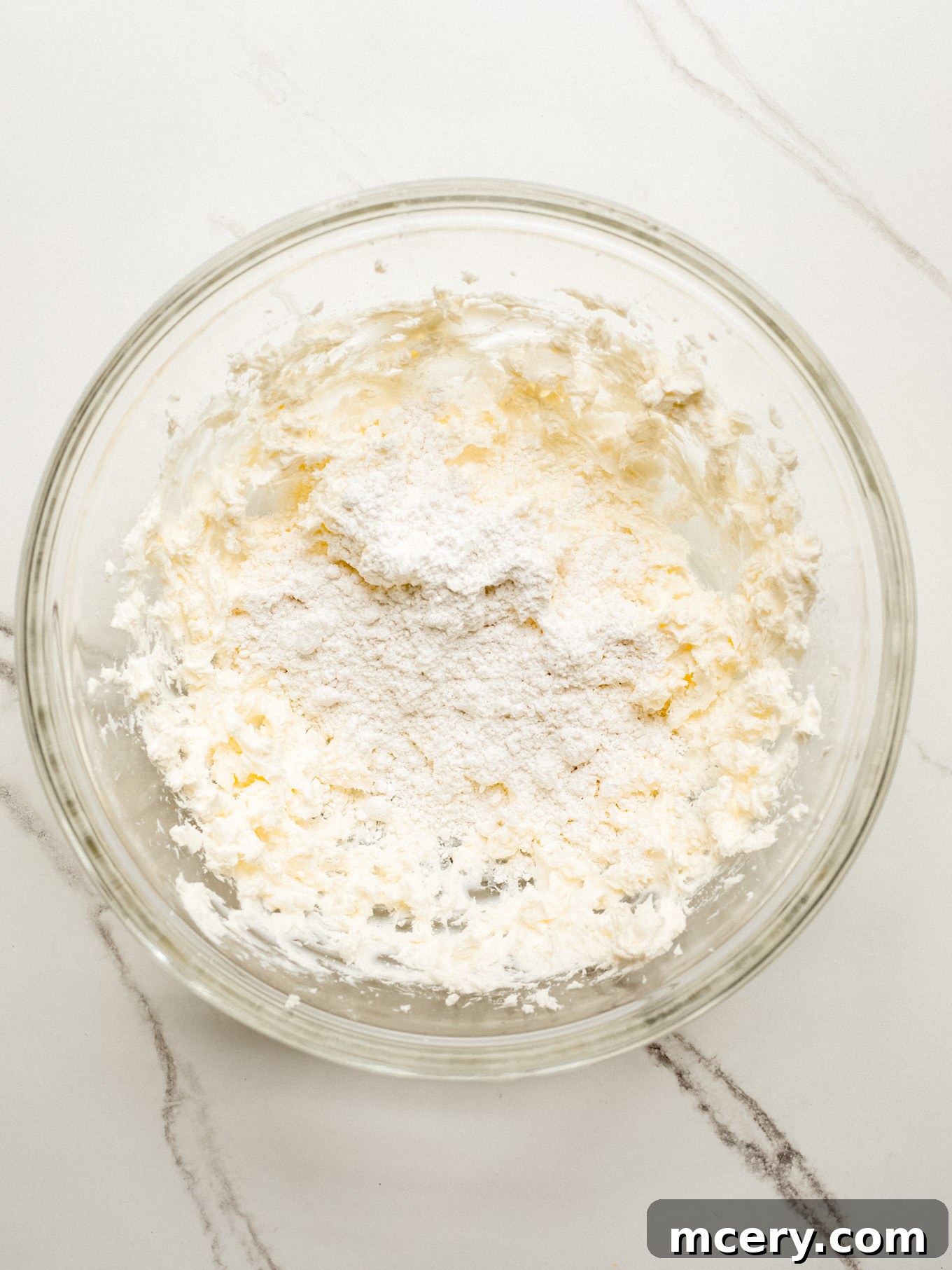 A bowl containing a creamy mixture of whipped cream cheese and powdered sugar, with a hand mixer ready to blend.
