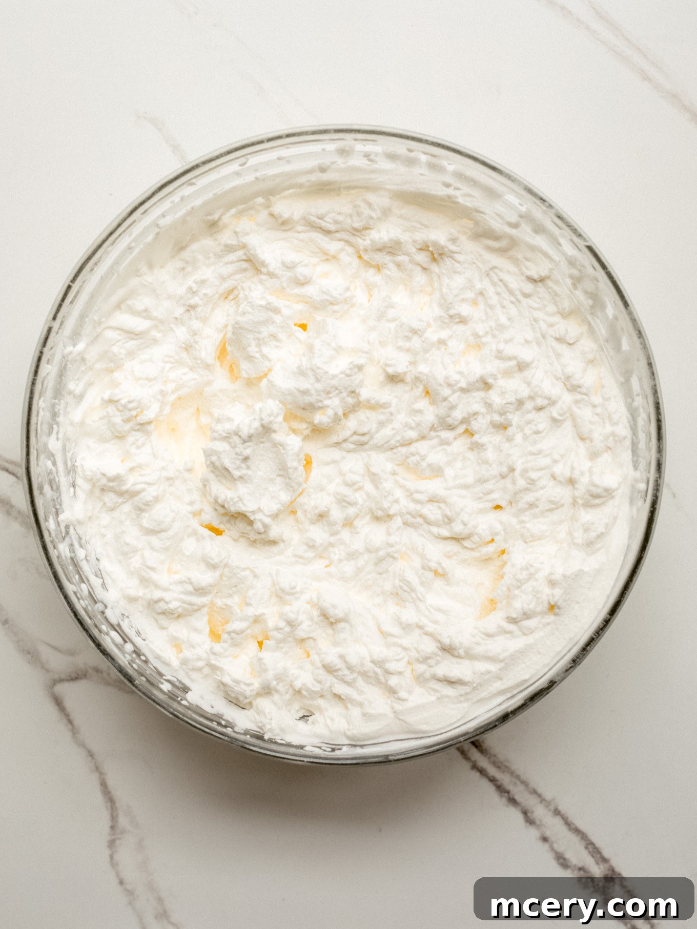 A clear glass mixing bowl filled with perfectly whipped heavy cream, showing stiff peaks that indicate it's ready for the next stage of the recipe.