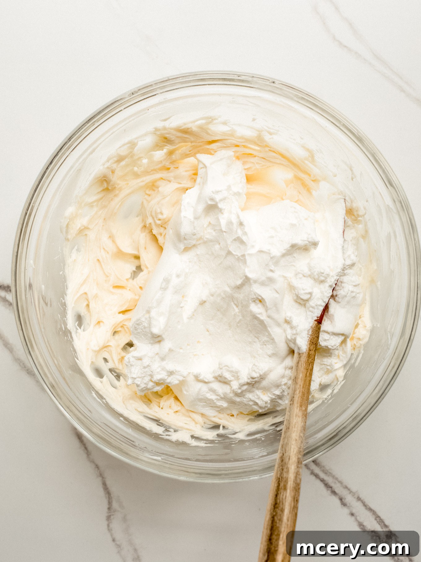 A rubber spatula gently folding the whipped cream into the cream cheese mixture in a glass bowl, creating a light and airy filling.