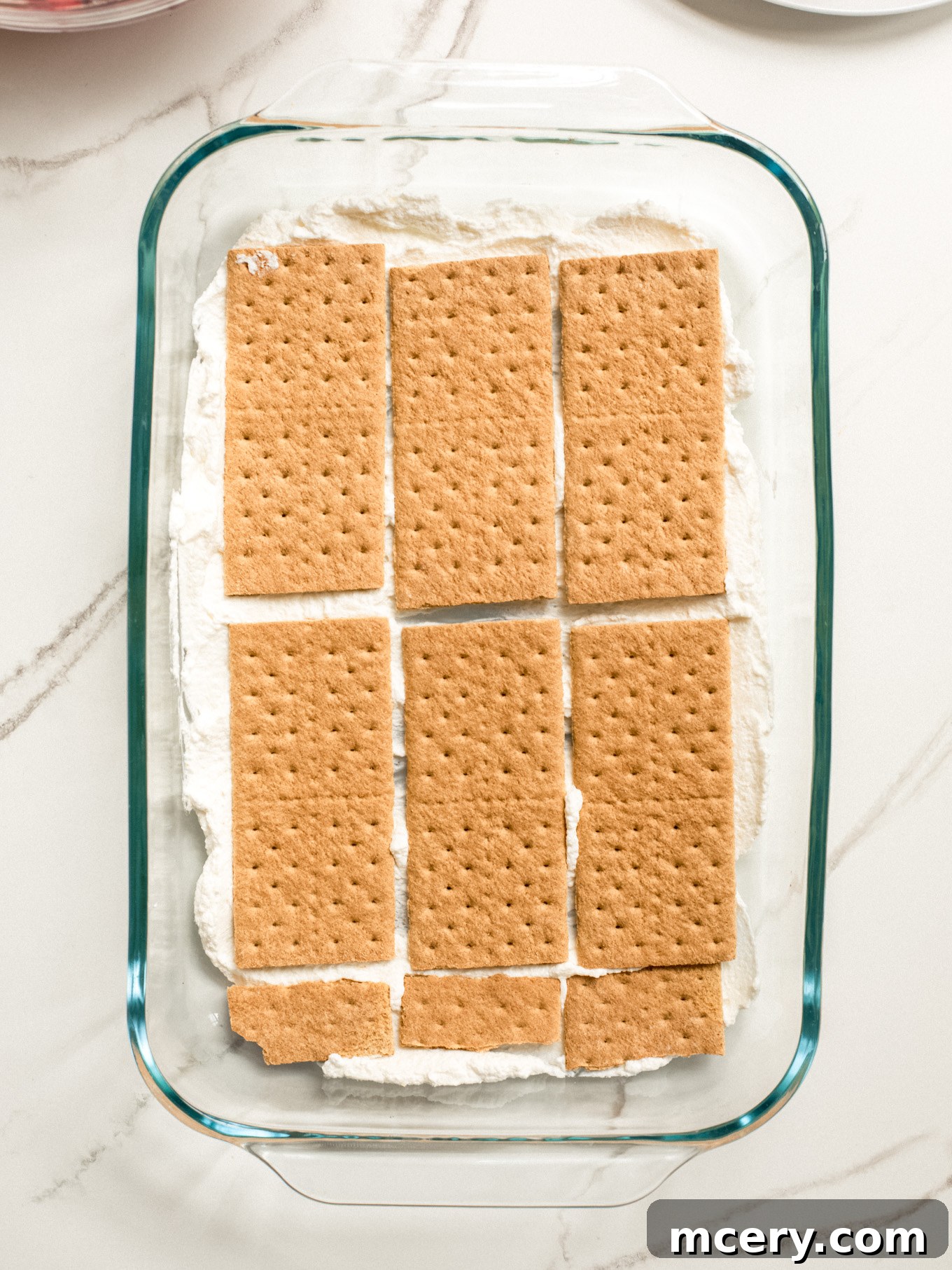The first layer of cream mixture thinly spread on the bottom of a glass baking dish, topped with a neat layer of graham crackers, serving as the base for the icebox cake.