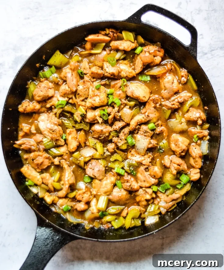 A skillet filled with a savory Black Pepper Chicken, featuring cooked pieces of chicken, diced onions, green bell peppers, and carrots. The dish is garnished with chopped green onions and seasoned with black pepper.