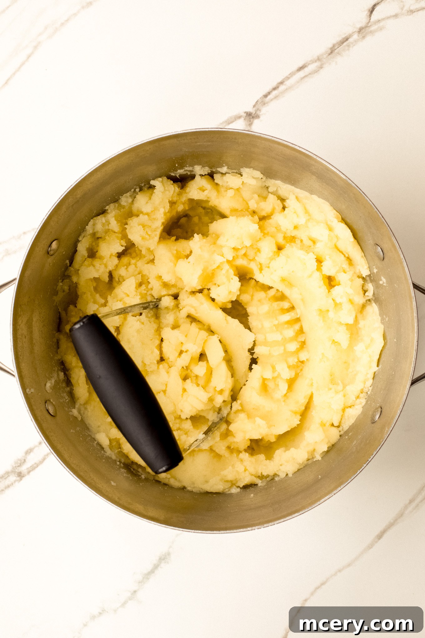 Silky Yukon Gold Mash 6 Mashed potatoes being worked with a masher in a stockpot, creating a smooth texture.
