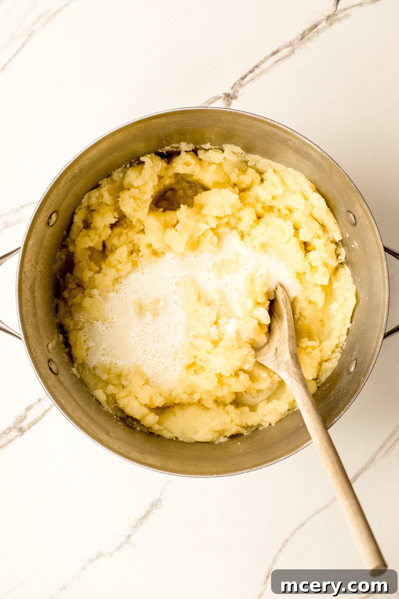 Silky Yukon Gold Mash 8 Mashed potatoes in a stockpot with half of the warm milk and butter mixture being folded in with a wooden spoon.