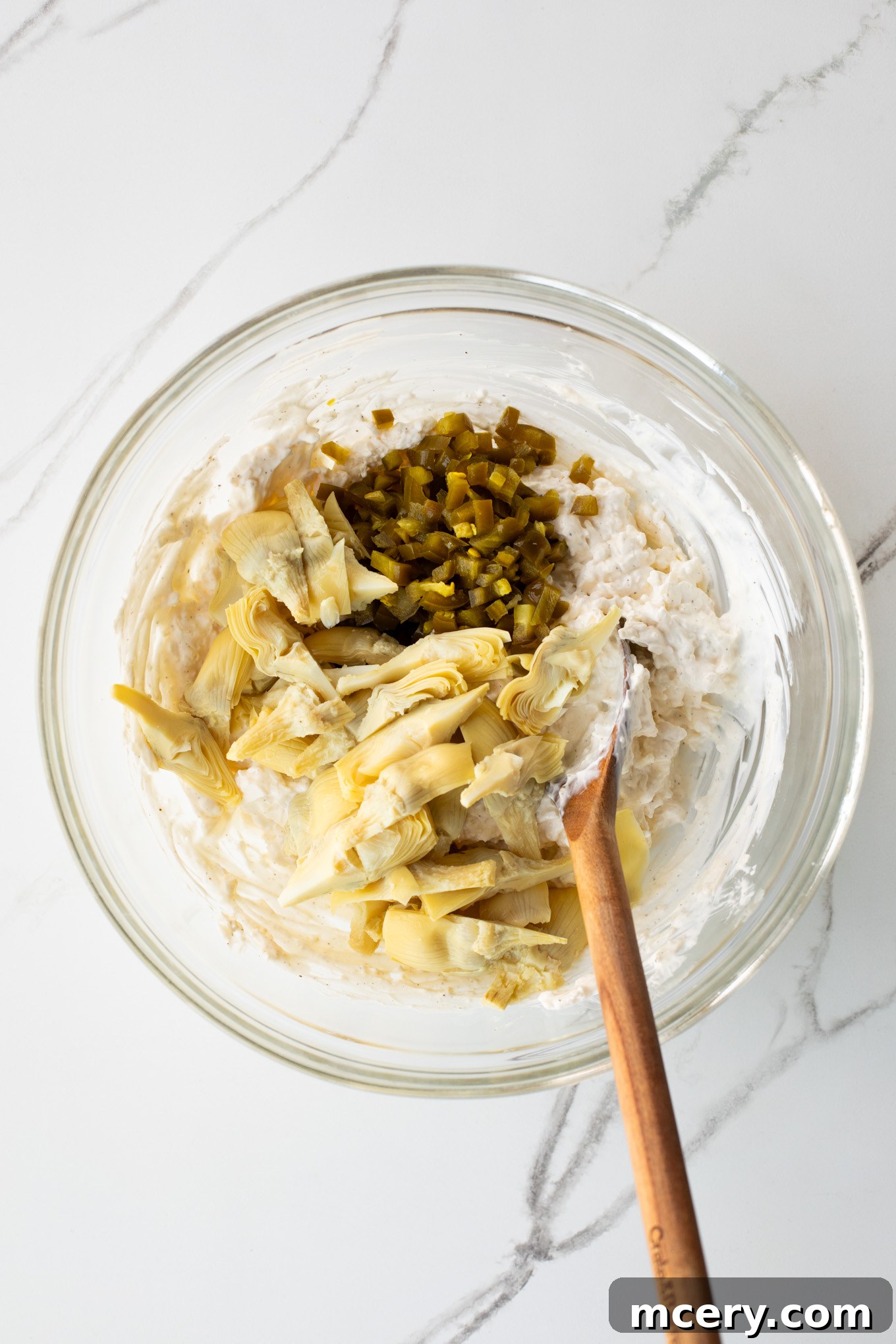 Spicy Jalapeno Artichoke Dip 6 Chopped artichoke hearts and pickled jalapenos are added to the creamy cheese mixture in a glass bowl.