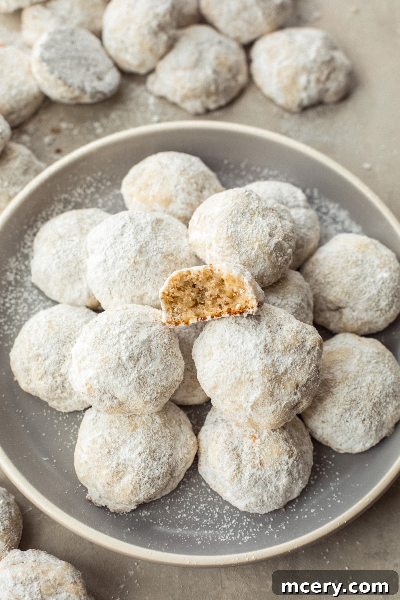Buttery Pecan Snowflakes 10 A beautifully arranged plate of pecan cookies, with one cookie delicately split in half to reveal its crumbly, nutty interior, all dusted with a generous layer of confectioners' sugar.