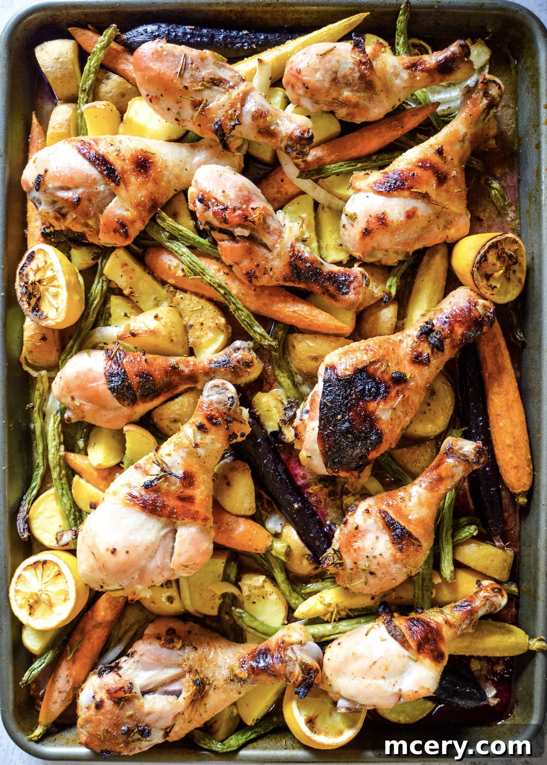 Golden brown garlic herb chicken drumsticks roasting on a sheet pan with colorful vegetables like carrots, potatoes, and green beans.
