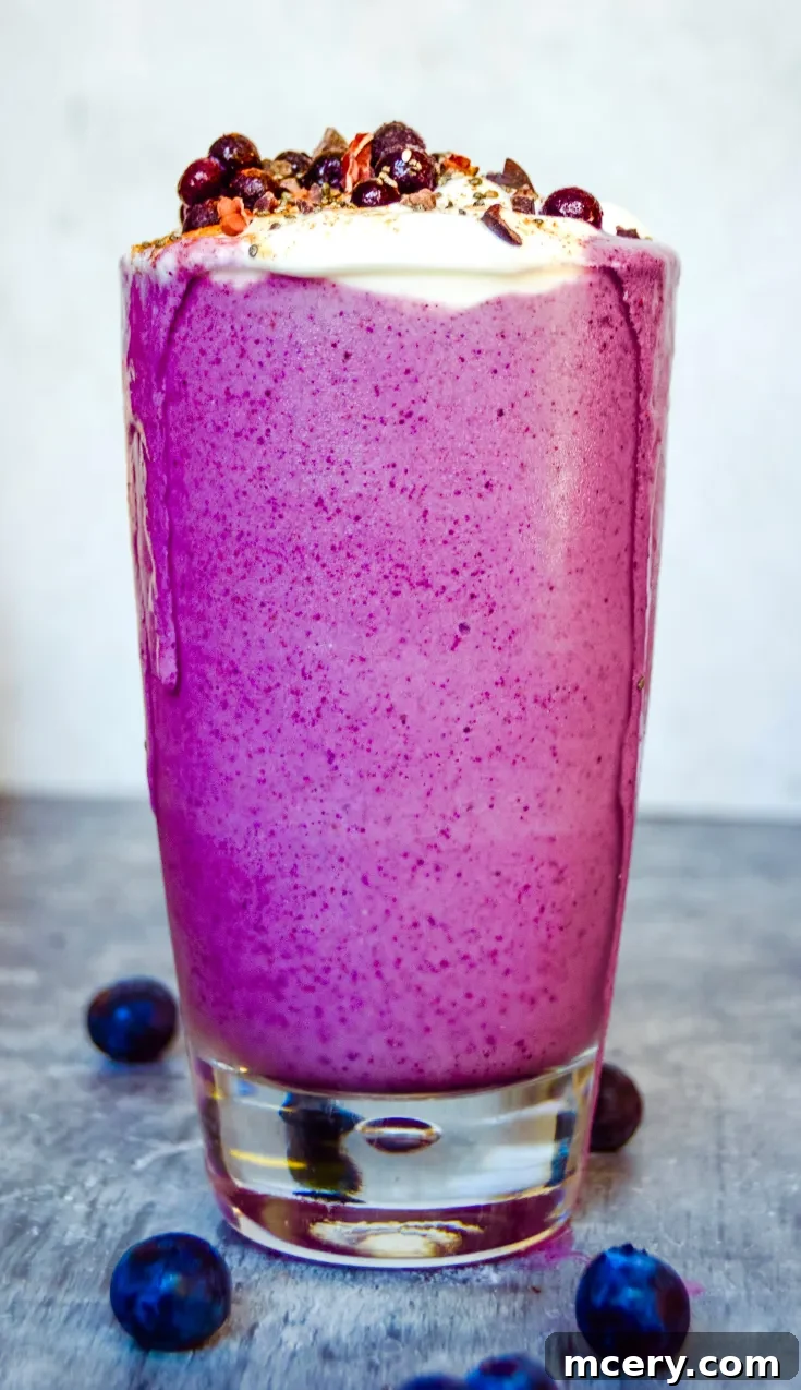 A tall glass of vibrant purple Pregnancy Blueberry Smoothie topped with whipped cream, sprinkled with chocolate shavings and berries. A jar in the background, set on a textured gray surface.