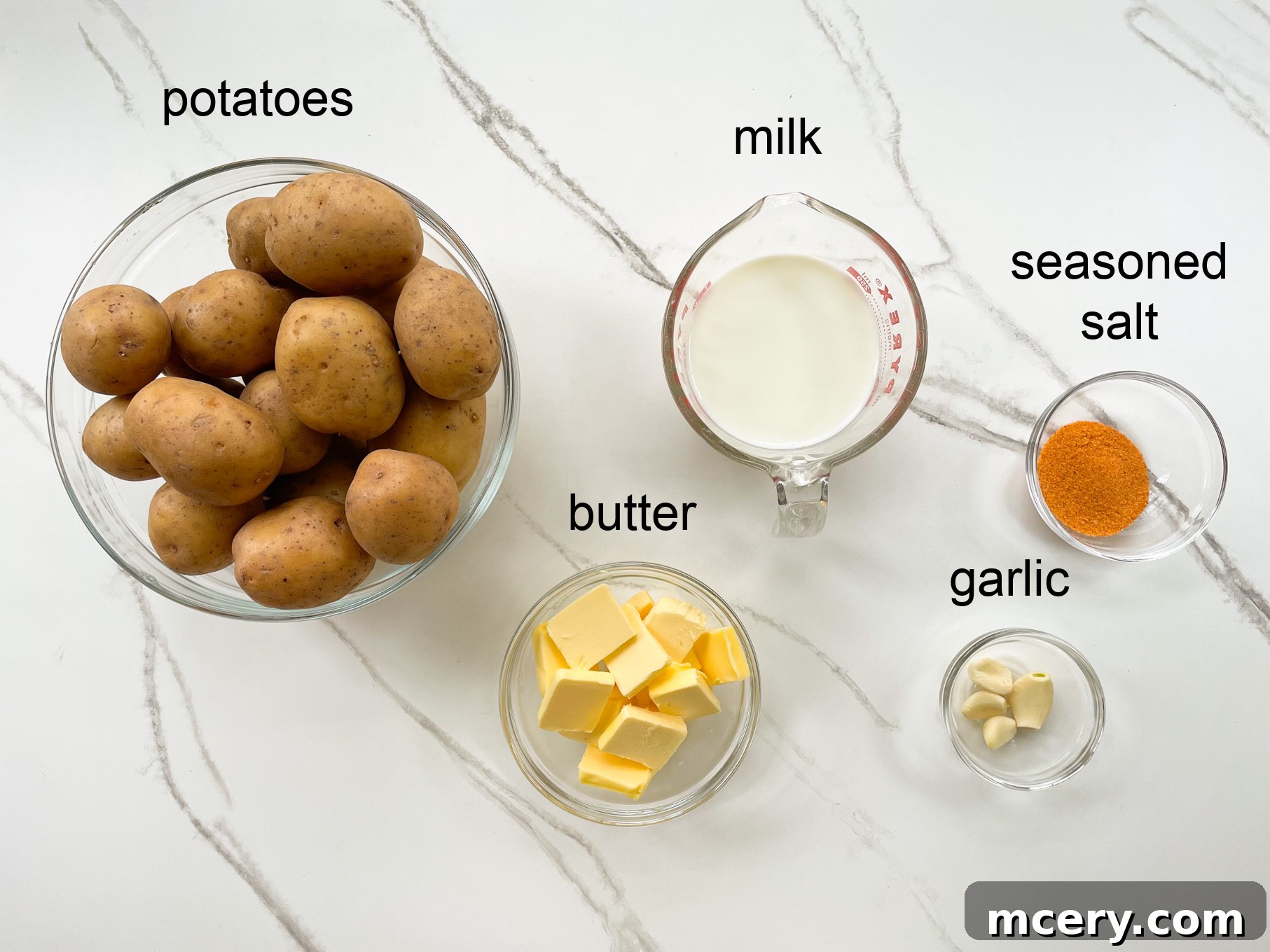 A collection of fresh ingredients laid out on a wooden board, including whole potatoes, a stick of butter, several cloves of garlic, and a small jug of milk, ready for making mashed potatoes.
