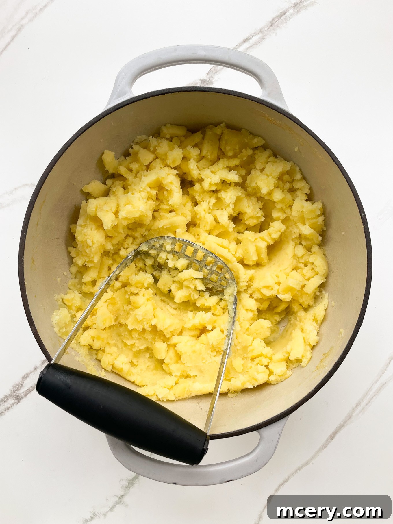 Freshly boiled and drained potatoes, partially mashed in a large cooking pot with a potato masher, awaiting the addition of dairy.