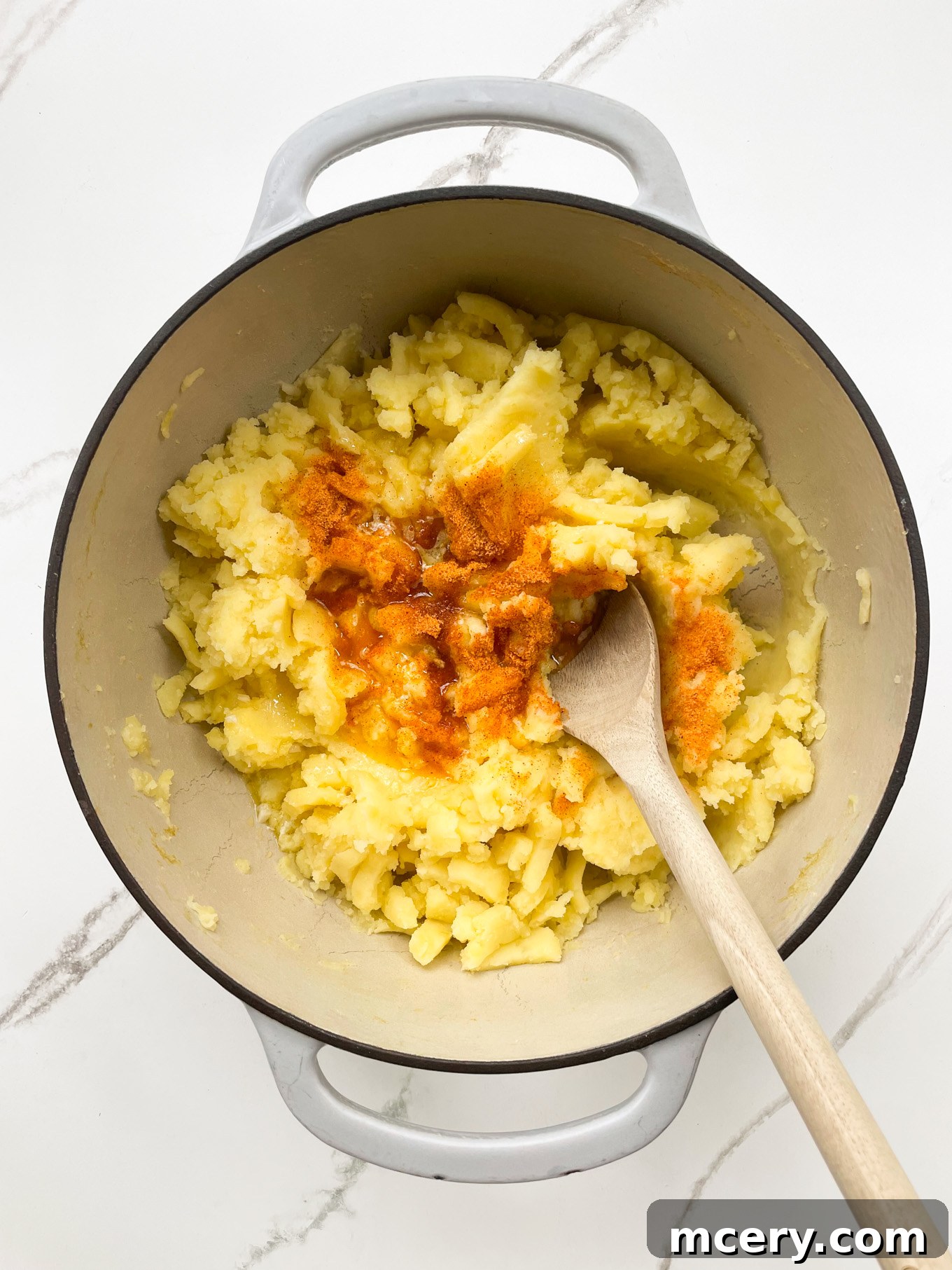 Half of the warm milk and butter mixture being poured over the mashed potatoes in a pot, along with a sprinkle of seasoned salt, ready for gentle folding.