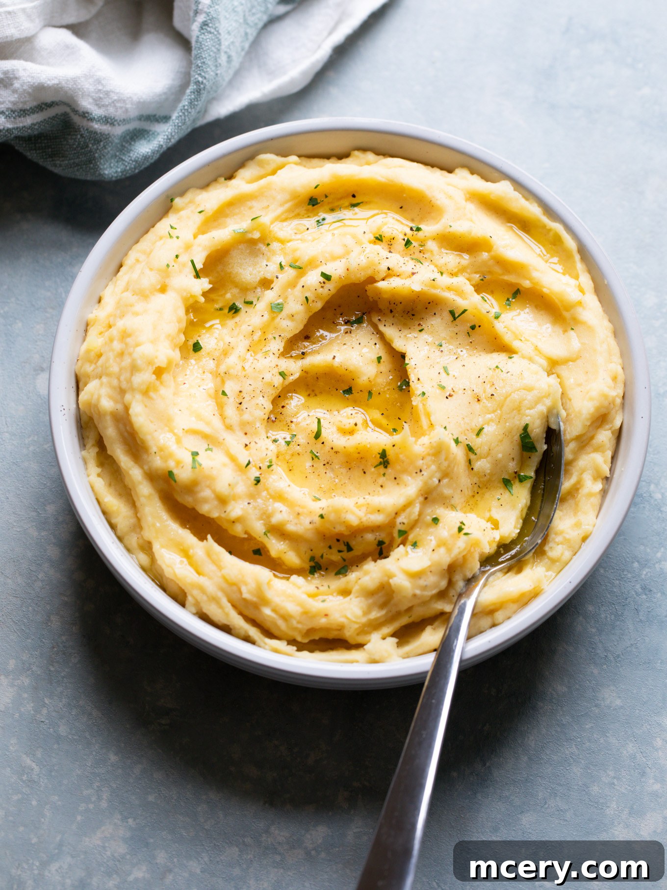 A serving of perfectly smooth and creamy garlic mashed potatoes presented in an elegant white bowl, accompanied by a silver serving spoon. The potatoes are garnished with fresh herbs.