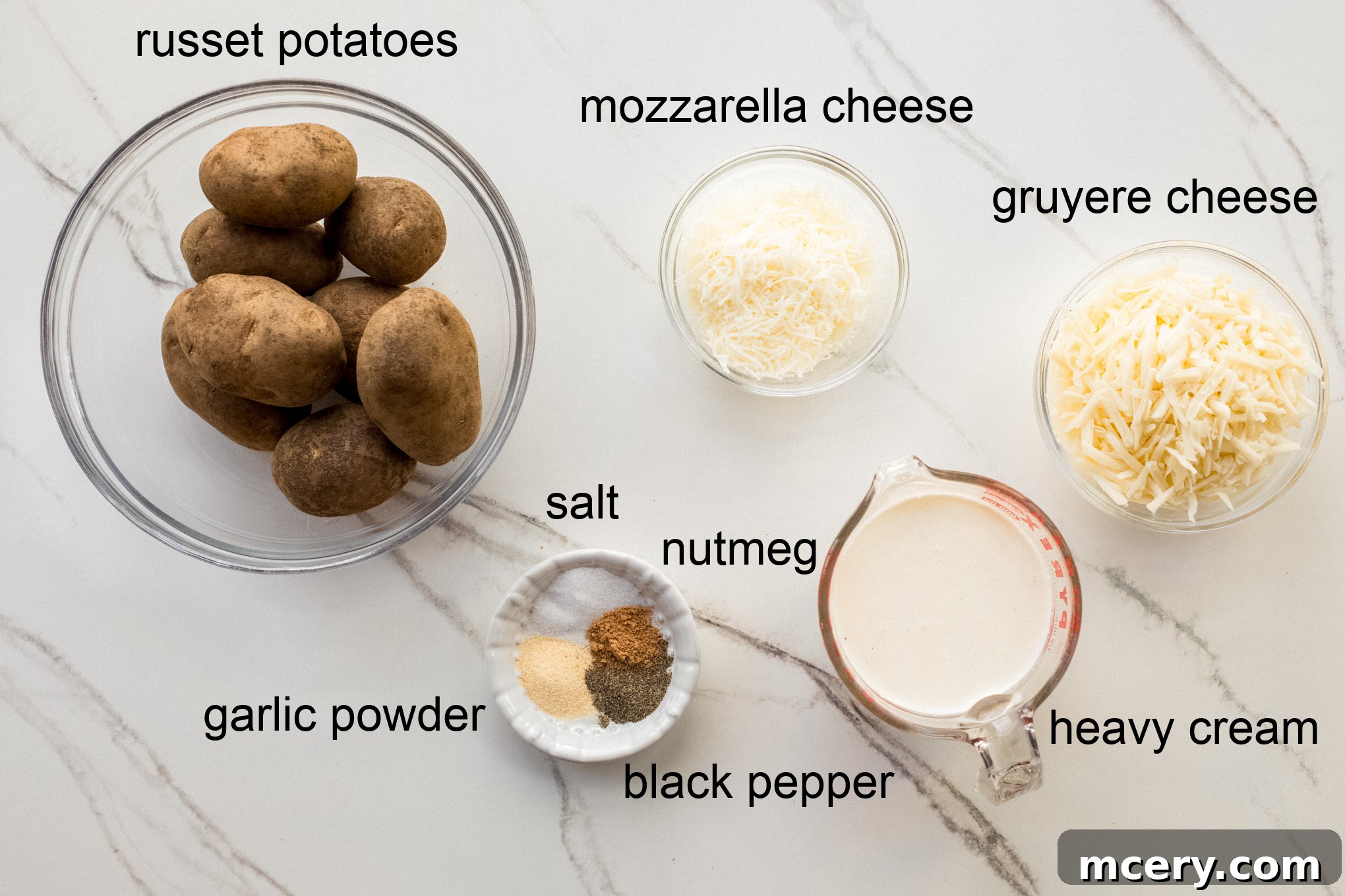 Decadent Cheesy Potato Gratin 3 A flat lay image showing all ingredients for making cheesy scalloped potatoes, including fresh potatoes, heavy cream, garlic powder, nutmeg, and two types of shredded cheese.