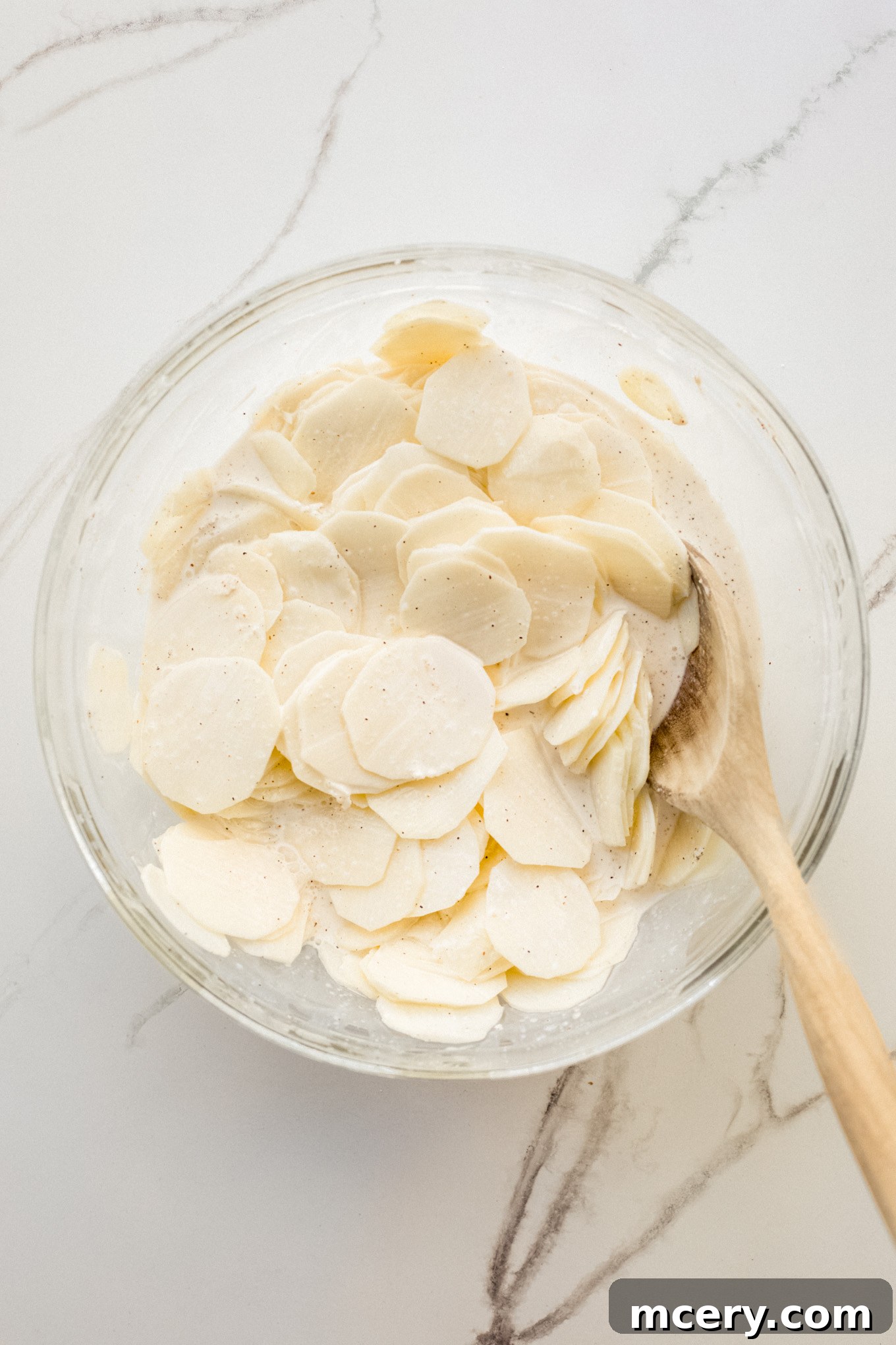 Decadent Cheesy Potato Gratin 5 Close-up of potato slices being thoroughly mixed with a wooden spoon in a bowl of seasoned cream, ensuring even coating.