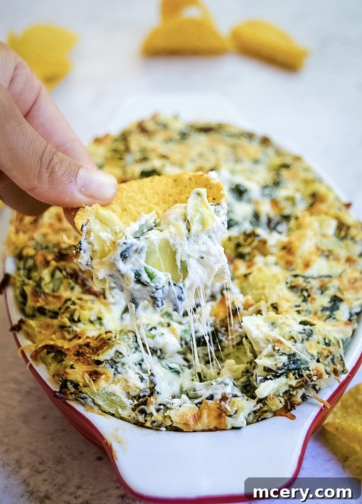 A hand dipping a round tortilla chip into a baked cheesy Spinach Artichoke Dip in a red-rimmed dish. The dip is creamy with visible chunks of artichoke and melted cheese strands stretching towards the chip.
