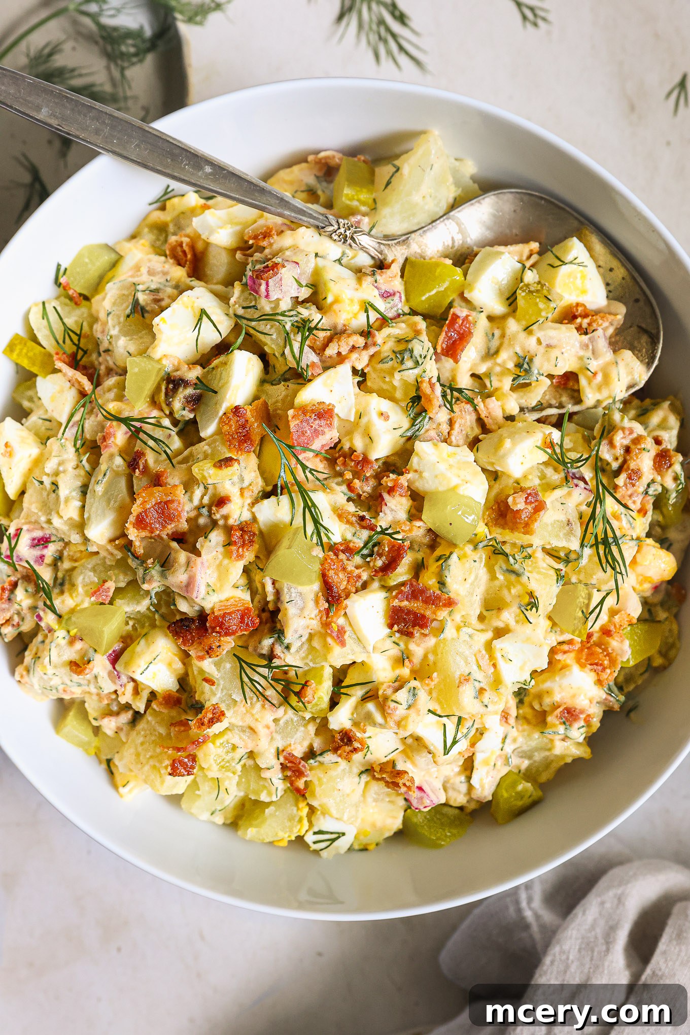 Close-up shot of creamy bacon potato salad in a white bowl, garnished with fresh dill and extra crispy bacon bits.