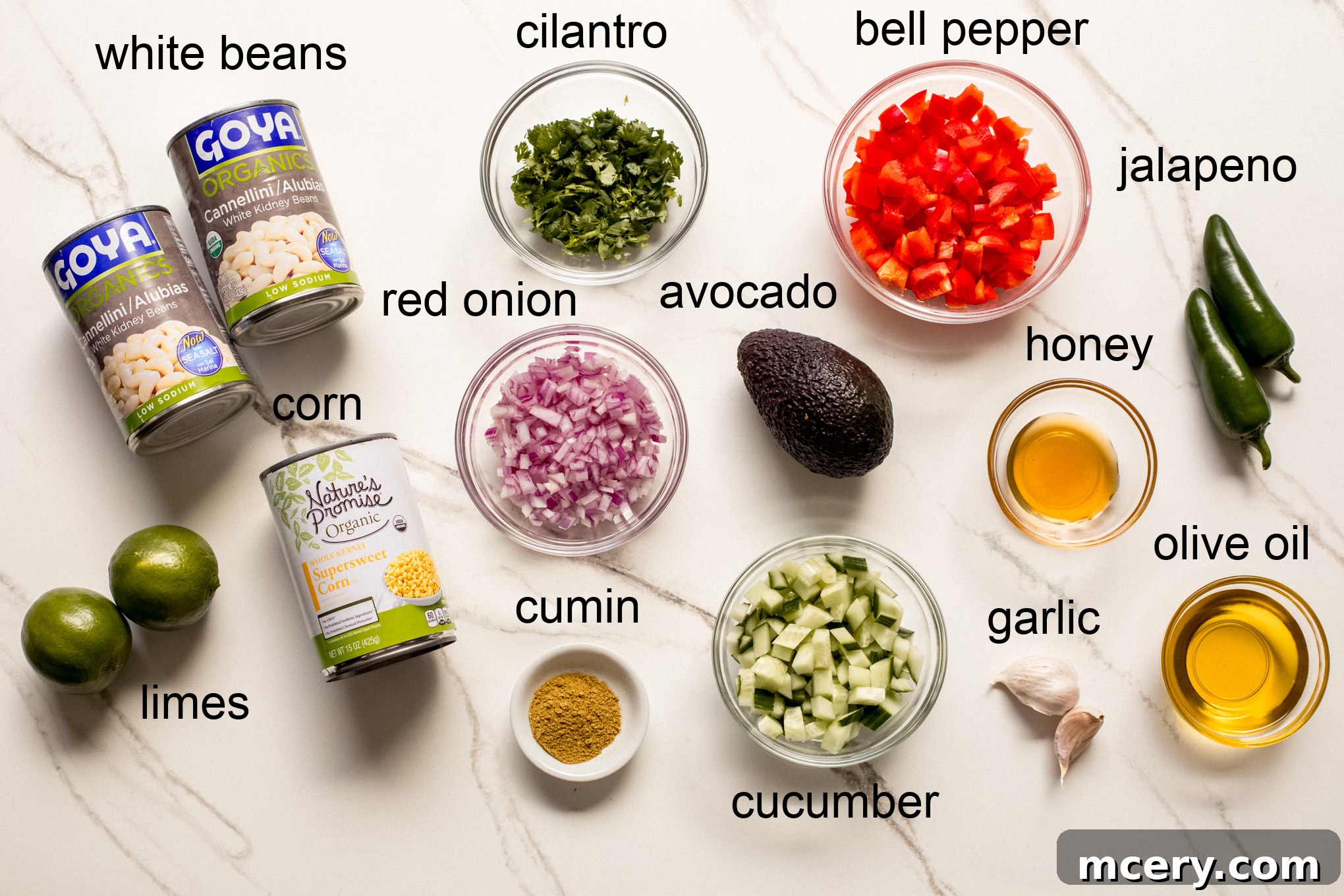 A selection of fresh ingredients laid out, including white beans, corn, cucumber, bell pepper, avocado, red onion, cilantro, and jalapeños.