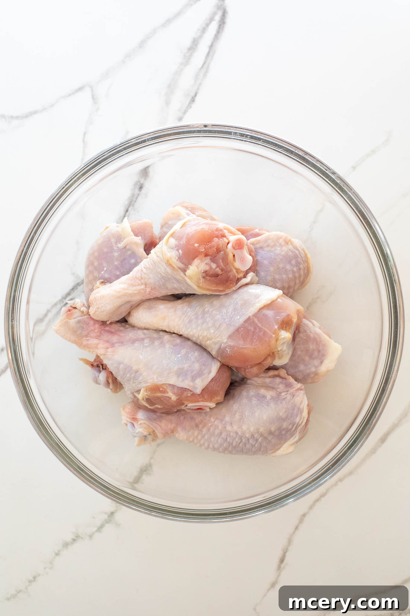 Sweet & Smoky Oven-Baked BBQ Drumsticks 4 Raw chicken legs in a clear glass bowl, glistening with oil.