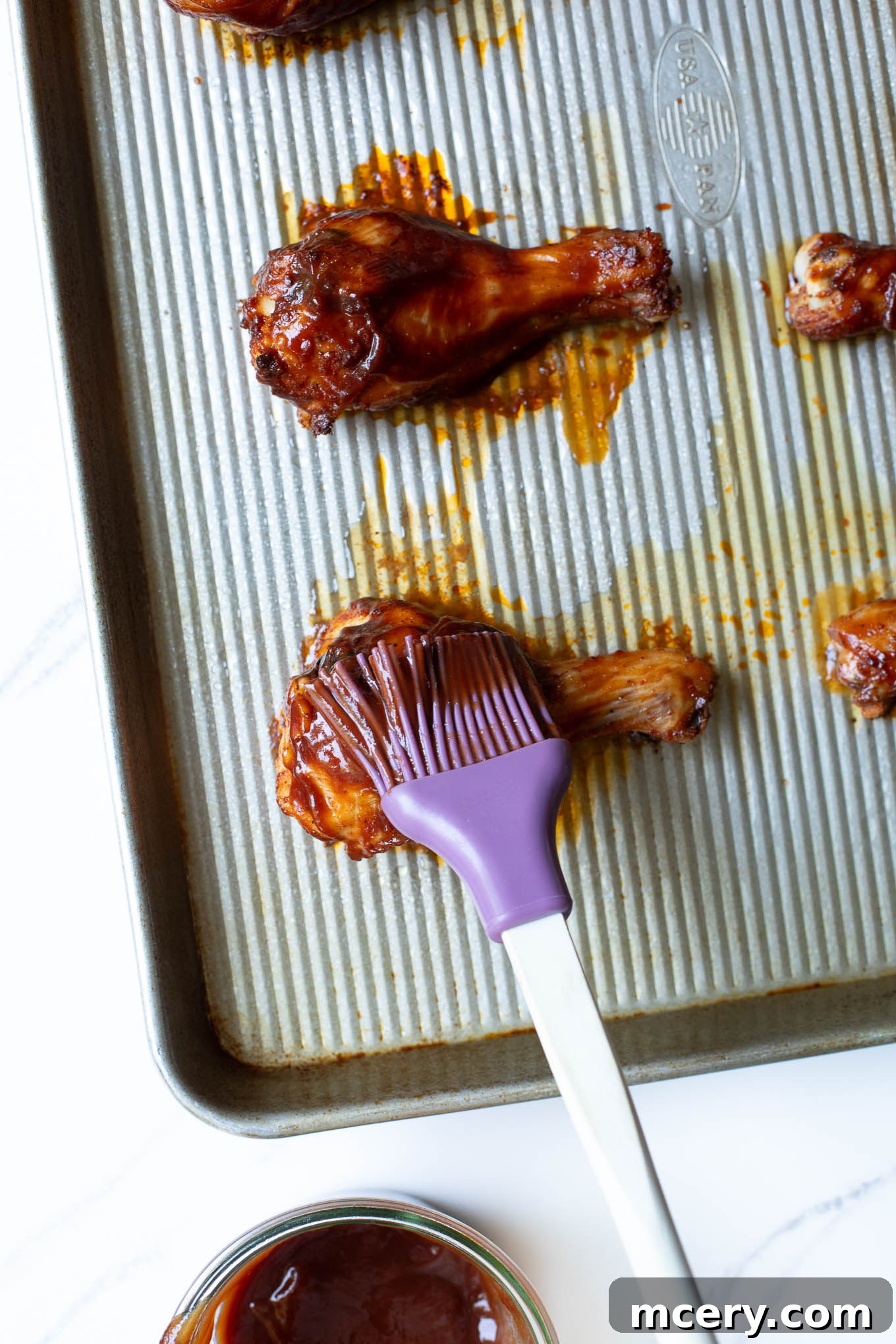 Sweet & Smoky Oven-Baked BBQ Drumsticks 8 A roasted drumstick being brushed with sticky barbecue sauce on a sheet pan.