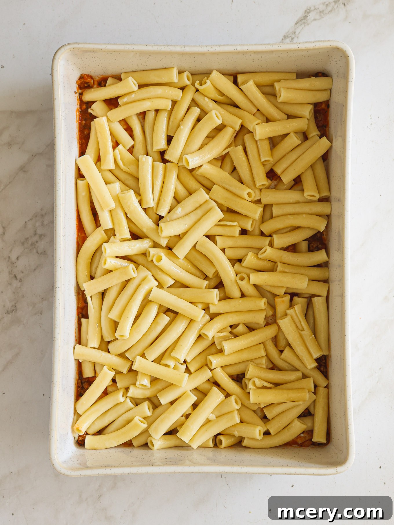 Golden Baked Ziti Delight 8 The first layer of baked ziti assembly: thin meat sauce topped with cooked ziti pasta in a baking dish.
