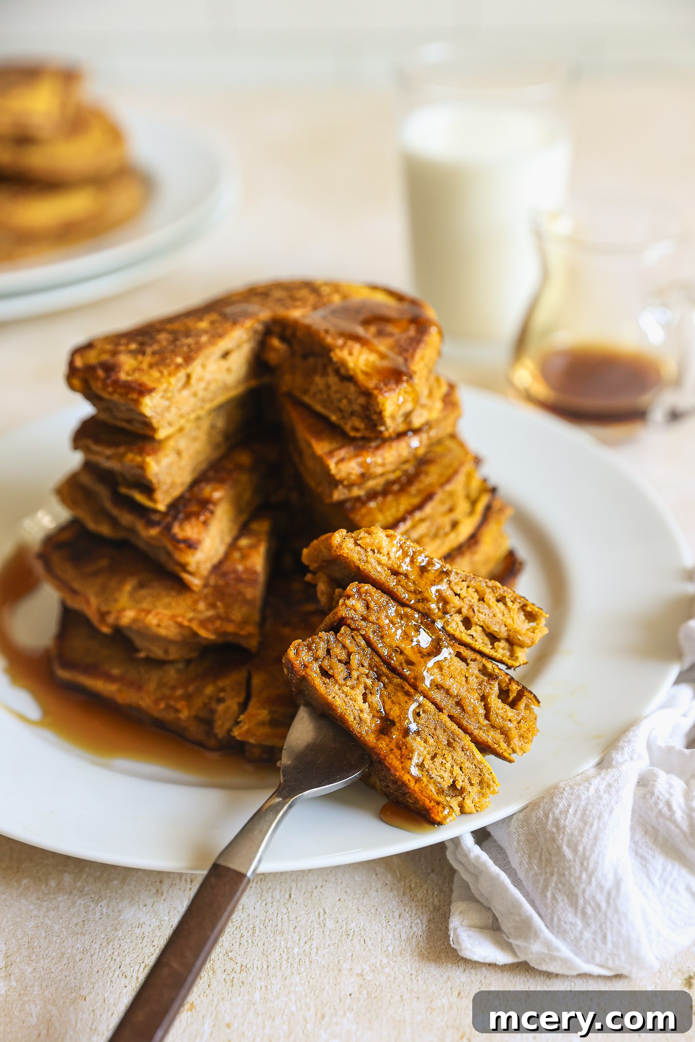 Whip Up Fluffy Pumpkin Pancakes 8 A stack of golden-brown pumpkin spice pancakes on a white plate, with one pancake cut open to reveal its fluffy interior. Drizzled with syrup.