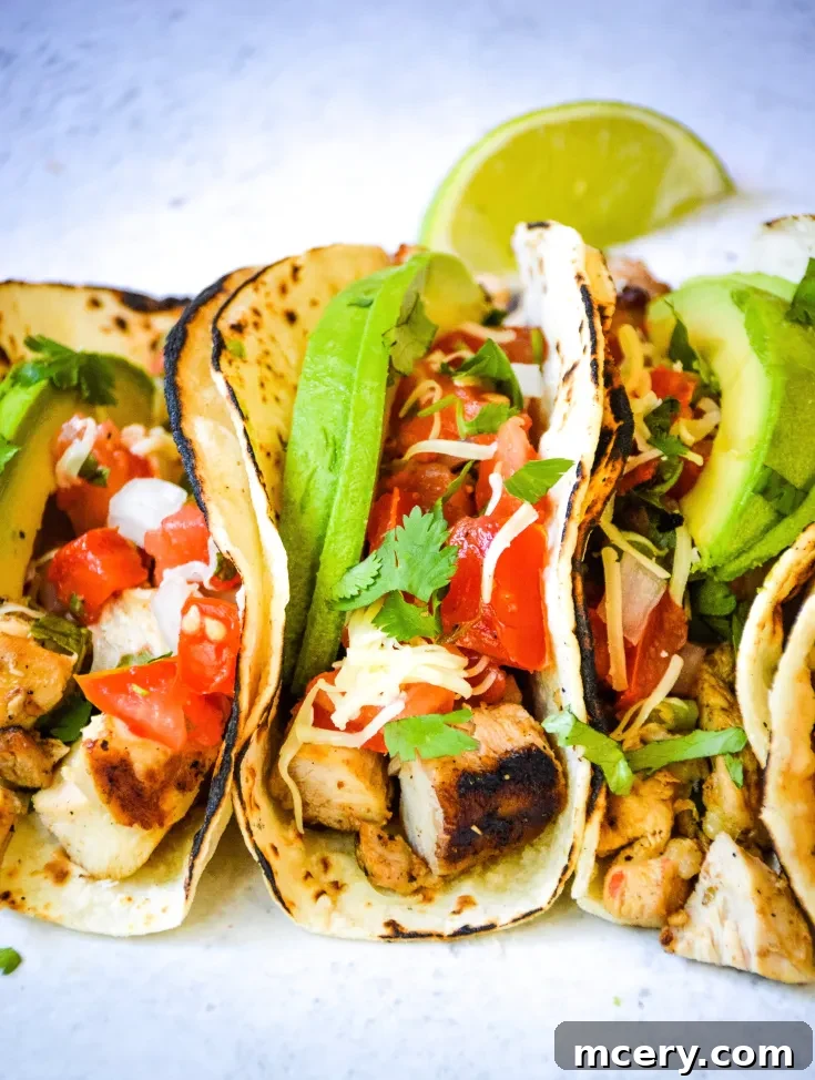 Three grilled Fresco Chicken Tacos on flour tortillas with diced tomatoes, shredded cheese, and cilantro. A lime wedge is on the side, placed on a light stone surface.