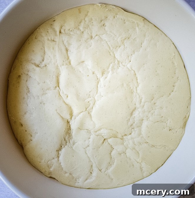 Fast and Easy Food Processor Pizza Dough 5 Pizza dough rising in an oiled glass bowl, covered with plastic wrap, showing its increased volume.