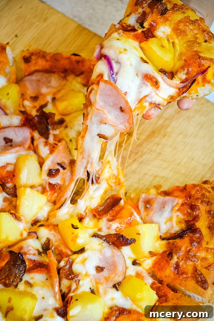 Close-up of a perfectly cooked slice of Hawaiian pizza being pulled from the whole pie, showcasing the gooey cheese and abundant toppings.