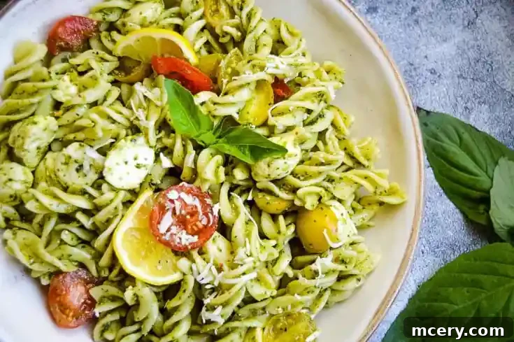 A bowl of fusilli Spinach Pesto Pasta Salad topped with halved cherry tomatoes, yellow lemon wedges, and fresh basil leaves. The pasta is coated in a green pesto sauce and sprinkled with grated cheese. Two large basil leaves are placed next to the bowl on a textured surface.