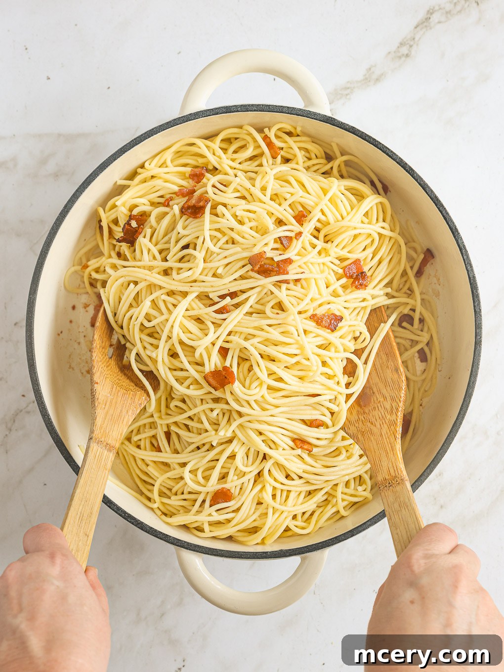 Hot cooked spaghetti being tossed with crispy bacon in a large skillet, coated in bacon fat.