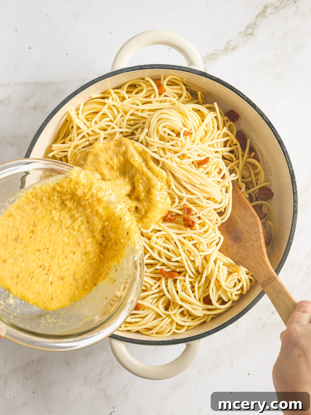 The egg and cheese mixture being quickly poured into the hot spaghetti and bacon in a skillet, stirred continuously.