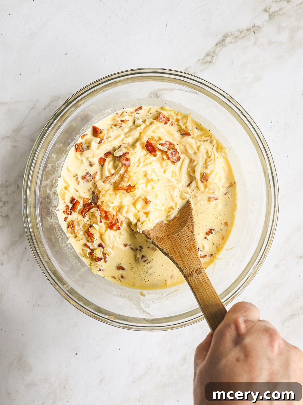 Efsane Hamursuz Kiş Tarifi 8 Whisked egg mixture with grated Swiss cheese, cooked bacon, and sautéed onions mixed in a glass bowl, ready for baking.