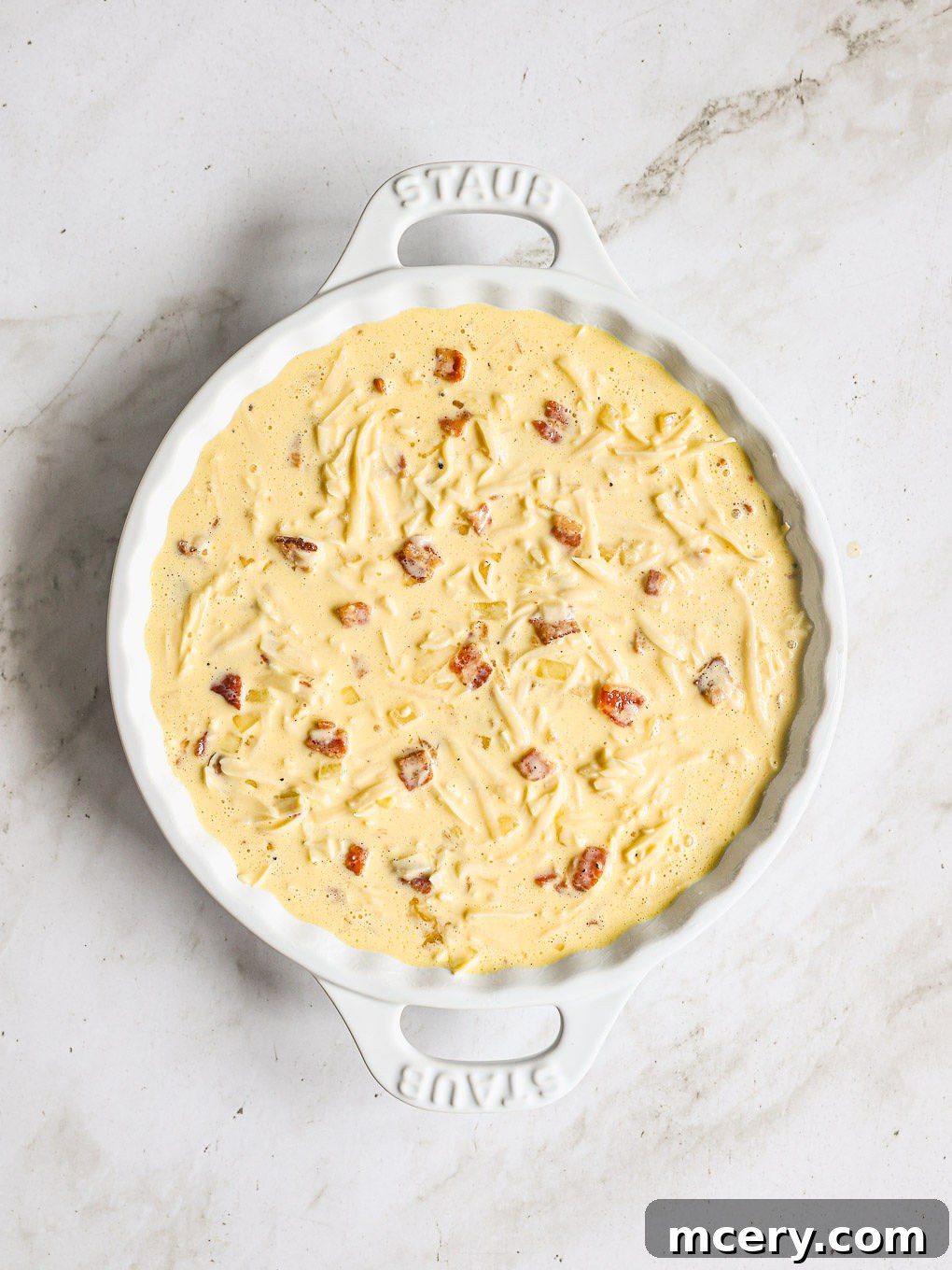 Efsane Hamursuz Kiş Tarifi 9 Quiche custard mixture poured into a greased baking dish, with ingredients spread evenly, ready to be baked.